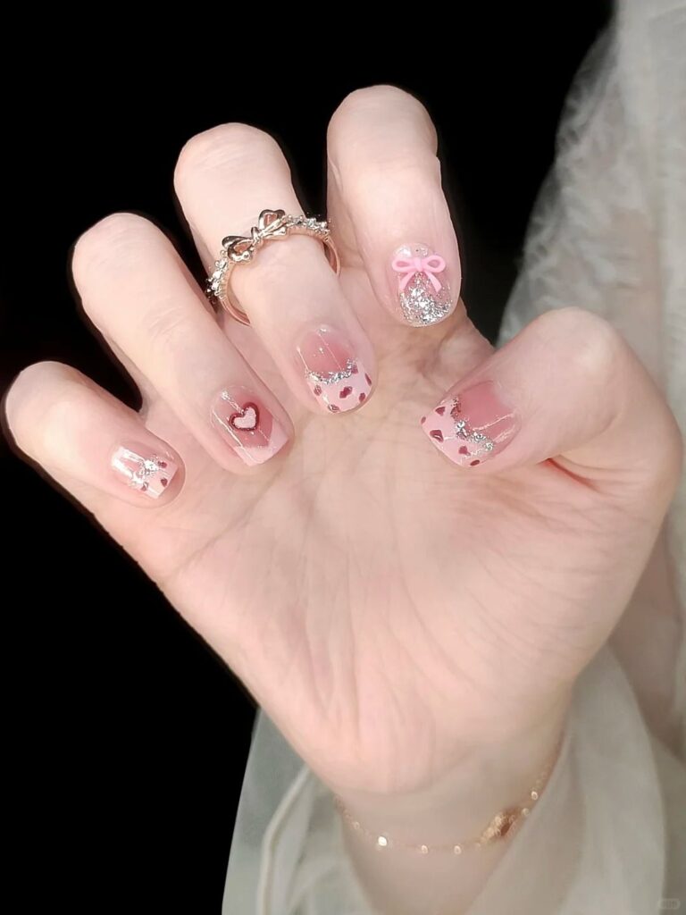 Mẫu nail cute cho móng ngắn nền hồng trong, họa tiết tim đỏ, nhũ bạc và nơ hồng 3D, ngọt ngào lãng mạn.