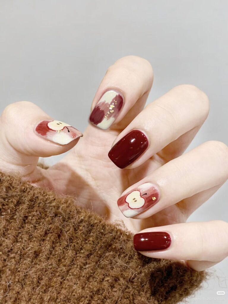 Mẫu nail đẹp tông đỏ mận bóng gương kết hợp nail ombre kem - đỏ vẽ họa tiết táo mini và nhũ vàng, phong cách ấm áp cho mùa thu đông.