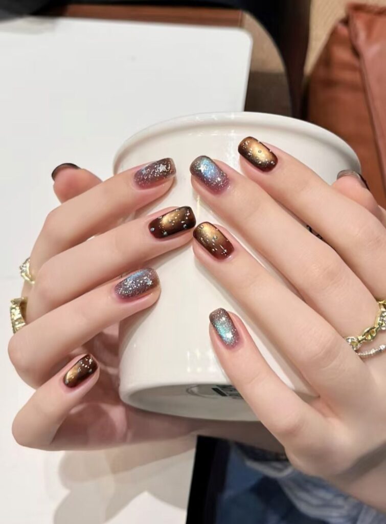 Mẫu nail mắt mèo nâu vàng ánh kim với họa tiết lấp lánh như dải ngân hà, điểm xuyết những chấm ánh sao nhỏ, sang trọng và cuốn hút.