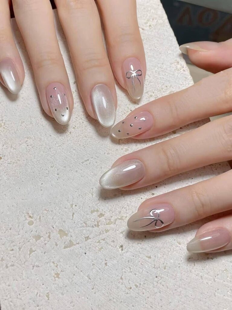 Mẫu nail mắt mèo trắng ngọc trai ánh kim lung linh, phối cùng móng nude đính đá nhỏ và họa tiết nơ ánh bạc vẽ tay tinh xảo, tinh khôi.