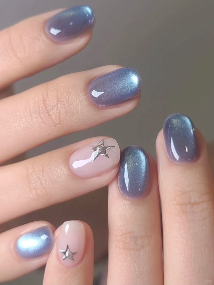 Mẫu nail mắt mèo xanh tím khói với hiệu ứng ánh kim huyền ảo đặc trưng, một móng nền nude đính charm ngôi sao bạc nhỏ xinh, nổi bật cá tính.