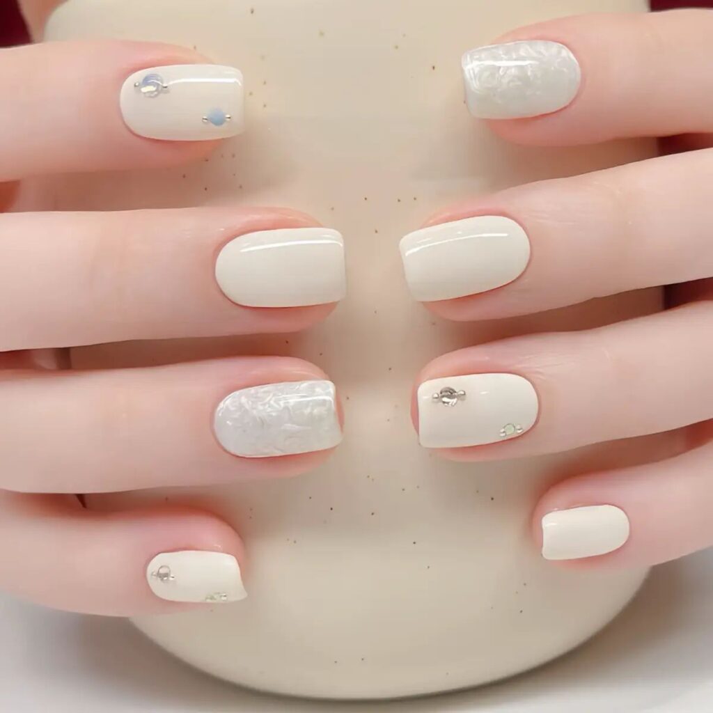 Nail màu trắng đơn giản dáng vuông nền trắng ngà bóng, điểm móng vân đá trắng ánh xà cừ cùng vài viên đá đính và hạt ánh xanh nhẹ nhàng.