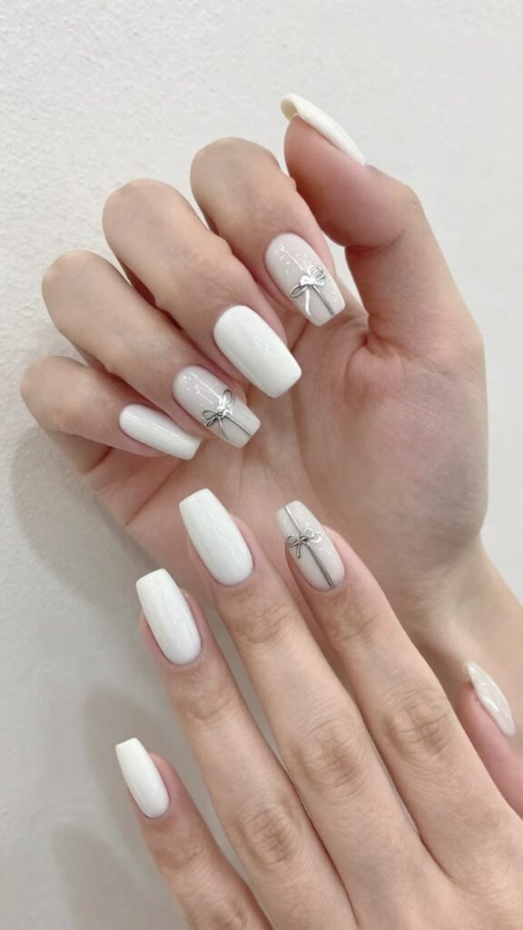 Nail màu trắng dáng coffin sơn trắng bóng, điểm móng nền sữa gắn nơ ánh bạc và kẻ chỉ bạc mảnh, nhẹ nhàng và tinh tế.
