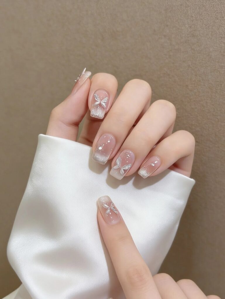 Những mẫu nail màu trắng đẹp nền nude trong, trang trí họa tiết bướm ánh bạc kim tuyến lấp lánh, nữ tính và tinh tế.